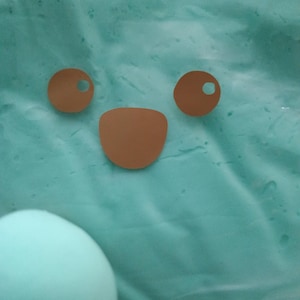 May include: A close-up of a light blue, textured surface with a cartoon face made of brown paper cutouts. The face includes two circular eyes with holes and a curved mouth. A light blue sphere is in the lower left corner.