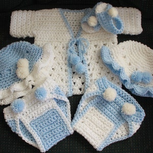 May include: A crocheted baby outfit set in white and light blue. The set includes a cardigan, a hat, booties, and a diaper cover. Each piece is decorated with white and blue pom-poms.
