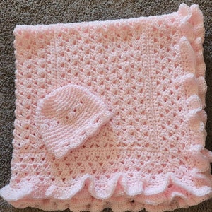 May include: A pink crocheted baby blanket with a matching crocheted hat. The blanket has a ruffled edge.