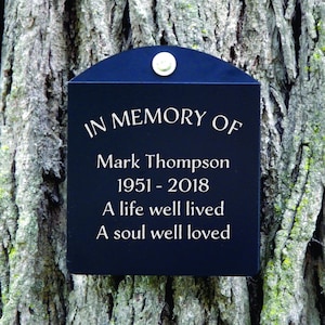May include: Black memorial plaque with rounded top edge attached to a tree trunk. The plaque reads "In Memory of Mark Thompson 1951-2018 A life well lived A soul well loved".