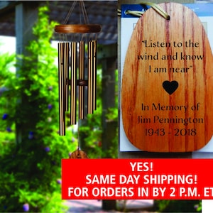 May include: Wooden wind chime with a heart and the text "Listen to the wind and know I am near" in memory of Jim Pennington 1943-2018.  The wind chime is hanging from a tree branch in front of a green leafy background.