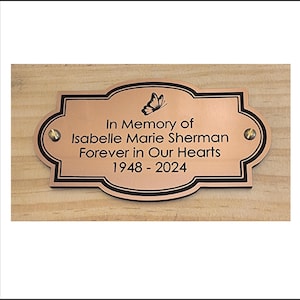 May include: A copper-colored memorial plaque with a black border and a butterfly design. The plaque reads "In Memory of Isabelle Marie Sherman Forever in Our Hearts 1948 - 2024".