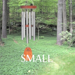 Pu&ograve; includere: Un carillon in argento con una cima in legno e un'etichetta in legno che dice "Mountain Living". Il carillon &egrave; appeso in un ambiente boschivo.