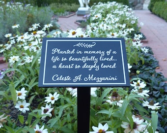 May include: A black plaque with white text reads "Planted in memory of a life so beautifully lived... a heart so deeply loved Celeste A. Mezzanini". The plaque is mounted on a black metal stake and surrounded by white flowers.
