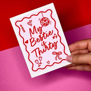 May include: A pink and white greeting card with red detailing. The card reads "My Bestie is Thirty" in a whimsical font, accompanied by illustrations of a disco ball, hearts, stars, a candle, and champagne glasses. The card is set against a red and pink backdrop.
