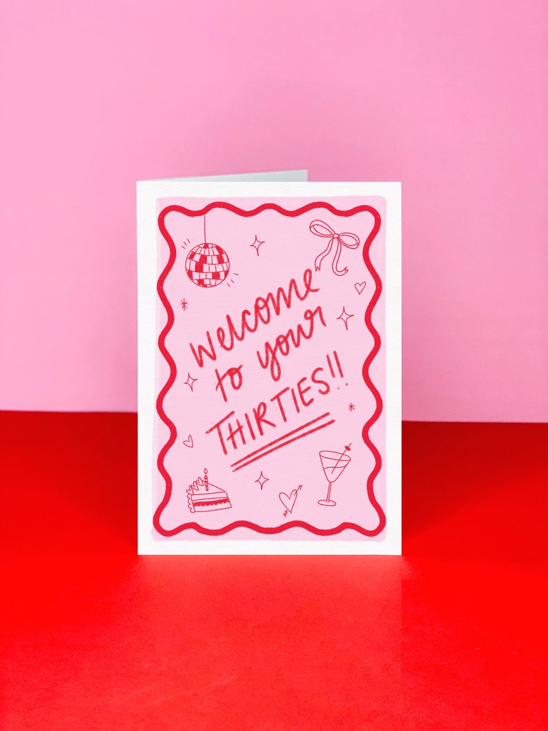 K&ouml;nnte beinhalten: Eine rosa Gru&szlig;karte mit wei&szlig;em Rand und rosa Innenleben. Die Karte hat einen welligen Rand und zeigt eine Discokugel, eine Schleife, einen Kuchen, ein Martini-Glas und den Text "Welcome to your Thirties!" in Rot.