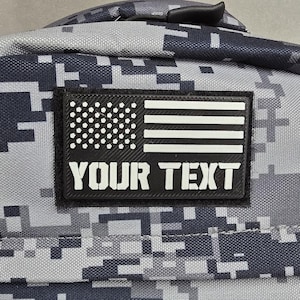 May include: A gray and blue digital camouflage backpack with a black patch featuring a white and black American flag design. The patch includes the text "YOUR TEXT" in white letters. The backpack has a zipper and a handle.