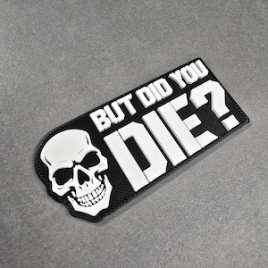 May include: A black rectangular patch with the phrase "BUT DID YOU DIE?" in white block letters. A white skull graphic is on the left side. The patch is on a gray surface.