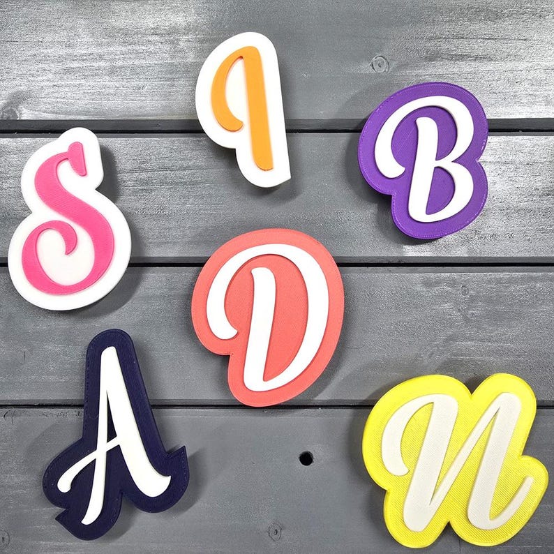 May include: Six colourful plastic cookie cutters shaped like the letters S, I, D, A, B, and N. The letters are in a cursive font and have a white outline.