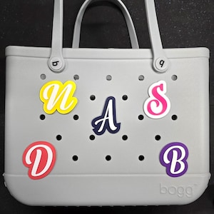 May include: Grey tote bag with a "BOGG" logo and colourful letters spelling "NASDB" attached. The letters are in various colours, including yellow, pink, blue, and purple.