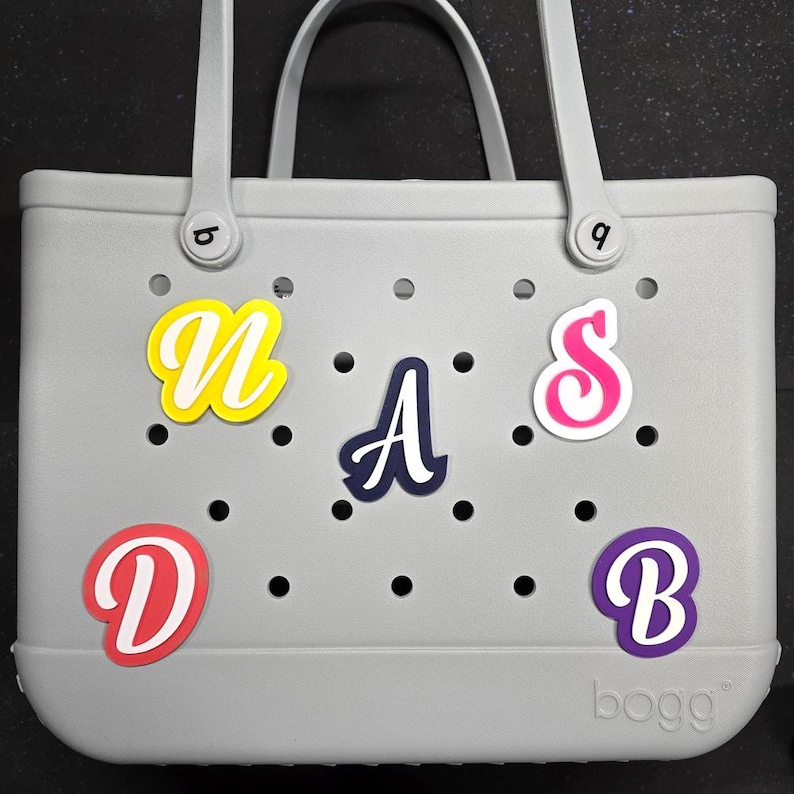 May include: Grey tote bag with a "BOGG" logo and colourful letters spelling "NASDB" attached. The letters are in various colours, including yellow, pink, blue, and purple.