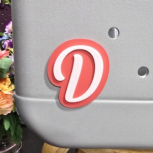 May include: A red and white letter D sticker. The letter is in a cursive font and is on a grey background.