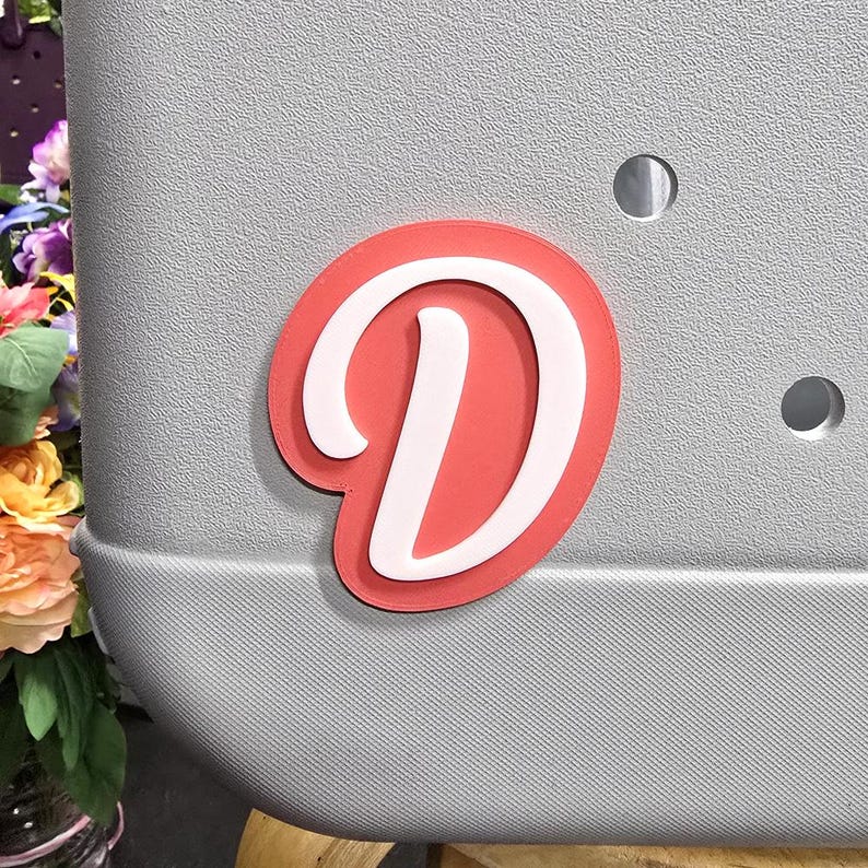 May include: A red and white letter D sticker. The letter is in a cursive font and is on a grey background.