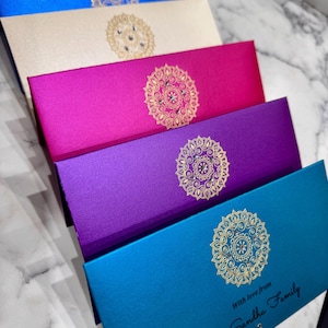May include: A stack of five rectangular envelopes in blue, cream, magenta, purple, and teal. Each envelope features a centred gold mandala design with small embedded crystals. The bottom teal envelope includes the black text With love from The Sandhu Family.