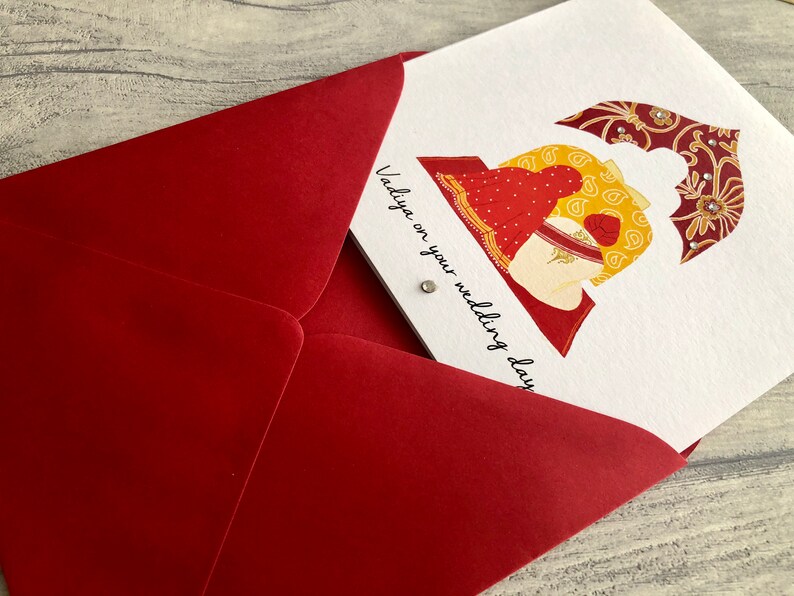 Anand Karaj Sikh wedding card is shown partially inside the red envelope. Illustration shows the Sikh couple sitting before the Guru Granth Sahib (Sikh Holy Book) Front is printed with, Vadiya on your wedding day.