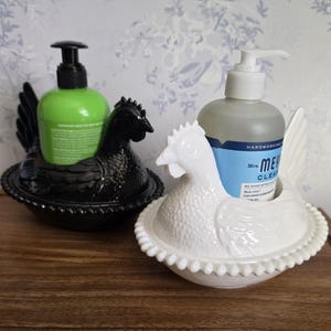 May include: Two ceramic chicken-shaped soap dispensers, one black and one white, each holding a bottle of liquid soap. The black chicken has a green bottle, and the white chicken has a clear bottle. Both dispensers sit on a wooden surface.