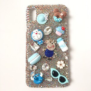 Puede incluir: Una funda de teléfono dorada brillante con adornos azules y blancos. La funda presenta una variedad de encantos, incluyendo un personaje de dibujos animados, una dona, un cupcake, una flor, gafas de sol y una botella de leche.