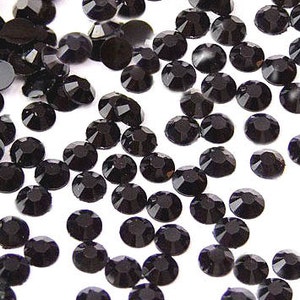 May include: A scattering of small, black, faceted rhinestones. Each gem has a round shape with multiple facets, creating a sparkling effect. The rhinestones are scattered across a white surface, with varying degrees of light reflection.