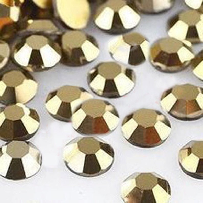 May include: A large quantity of gold-colored rhinestones, each with a faceted design. The rhinestones are scattered on a white surface.