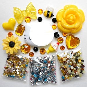 Könnte beinhalten: Ein Set mit gelben und weißen Verzierungen zum Basteln, darunter eine Sonnenblume, eine Rose, eine Schleife, eine Biene, ein Hello-Kitty-Gesicht und verschiedene Strasssteine und Perlen.