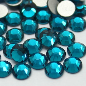 May include: A collection of teal blue rhinestones, also known as glass gems, scattered on a white surface. The rhinestones have a faceted design and a shiny, reflective finish.