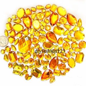 Pode incluir: Um grande sortimento de strass de vidro de cor âmbar em vários formatos e tamanhos. Os strass estão espalhados sobre um fundo branco. diyfashion123 está impresso na imagem.