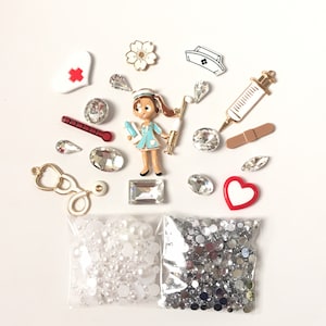 Puede incluir: Una colección de adornos blancos, plateados y rojos, que incluye una figura de enfermera, un corazón, una jeringa, un termómetro y una variedad de diamantes de imitación. Los adornos están dispuestos sobre una superficie blanca.