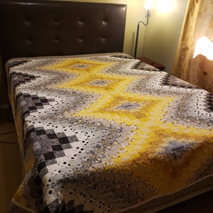 May include: A handmade quilt with a geometric pattern in shades of yellow, gray, black, and white. The quilt is spread across a bed with a brown leather headboard. The quilt's design features a central yellow stripe.
