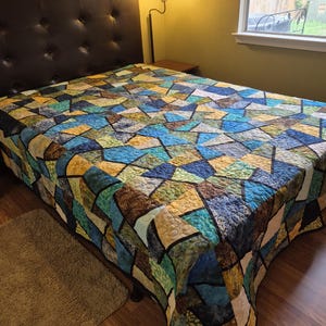 Puede incluir: Una colcha colorida con un patrón geométrico de formas azules, verdes, marrones y amarillas cubre una cama. La cama tiene un cabecero de cuero marrón oscuro. La habitación tiene un ambiente cálido y acogedor.