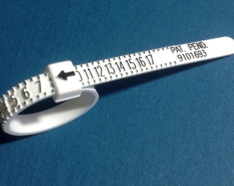 Könnte beinhalten: Ein weißer Kunststoff-Ringgrößenmesser mit einem schwarzen Pfeil und Zahlen von 4 bis 17. Der Text "PAT. PEND. 9101693" ist auf dem Größenmesser aufgedruckt.