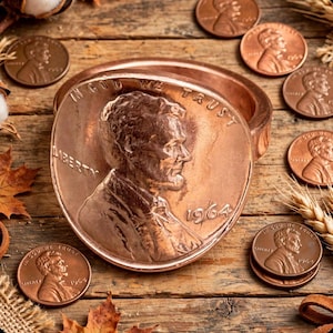 May include: A copper-colored circular container with a lid featuring the embossed profile of Abraham Lincoln, the text "IN GOD WE TRUST," "LIBERTY," and the year "1964." Several loose copper pennies are scattered around the central object.