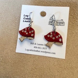 May include: A pair of mushroom-shaped earrings. Each earring features a red cap with white spots and a beige stem. The earrings are displayed on a white card with the text "Log Cabin Leather".