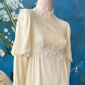 May include: Cream-colored vintage wedding dress with long sleeves and a high neckline, adorned with delicate lace detailing. The dress features a fitted bodice and a flowing skirt, displayed on a mannequin. A matching veil is also visible.