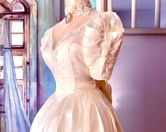 May include: A cream-colored satin gown featuring a V-neckline with lace trim, short puffed sleeves with horizontal pleats, and a full-length skirt. The dress includes a high lace choker collar with a small oval brooch at the center of the neck.
