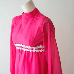 May include: A long-sleeved pink dress with a white crocheted trim around the waist. The dress has a high neck and long sleeves with buttoned cuffs.