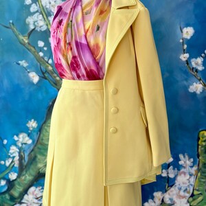 May include: A pale yellow skirt suit with a matching blazer and a floral scarf. The blazer has three buttons and a contrasting stitch detail. The skirt is pleated. The scarf has pink, purple, and yellow floral patterns.