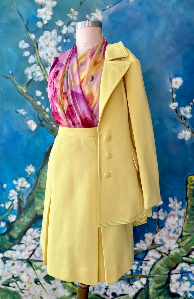 May include: A pale yellow skirt suit with a matching blazer and a floral scarf. The blazer has three buttons and a contrasting stitch detail. The skirt is pleated. The scarf has pink, purple, and yellow floral patterns.