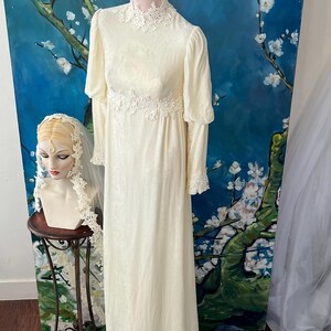 May include: Cream-colored vintage wedding dress with long sleeves and a high neckline. The dress features delicate lace detailing at the collar, waist, and cuffs. A mannequin displays the dress against a backdrop of a blue floral painting.