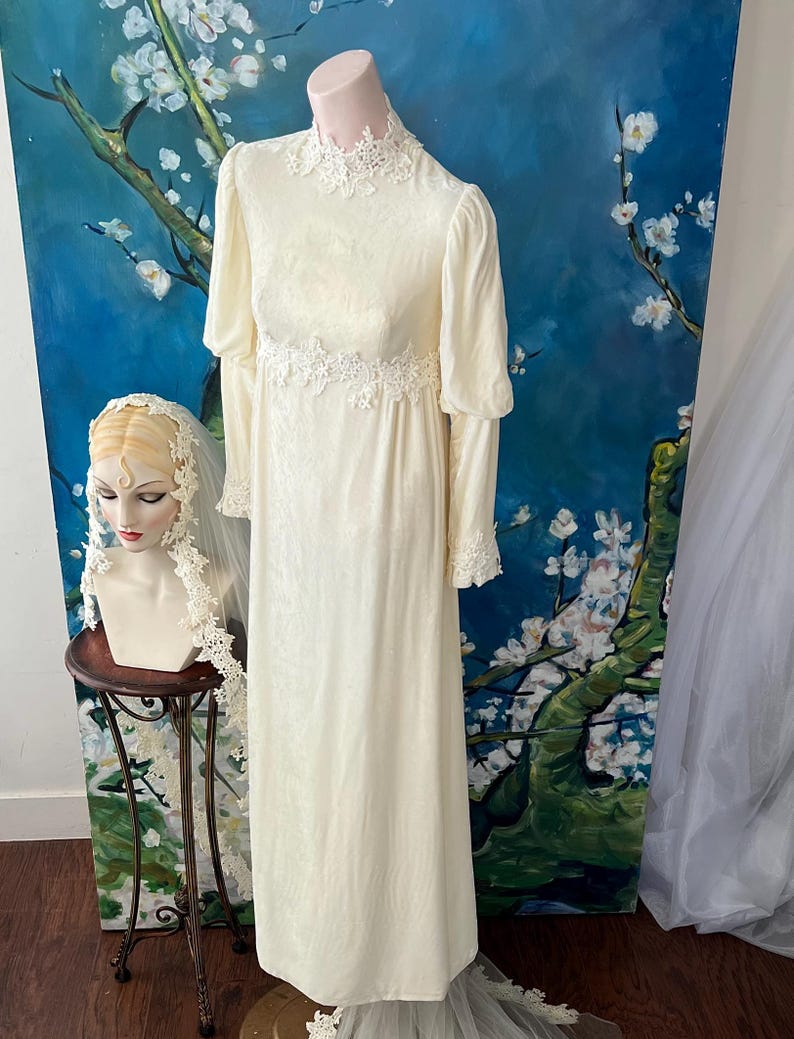 May include: Cream-colored vintage wedding dress with long sleeves and a high neckline. The dress features delicate lace detailing at the collar, waist, and cuffs. A mannequin displays the dress against a backdrop of a blue floral painting.