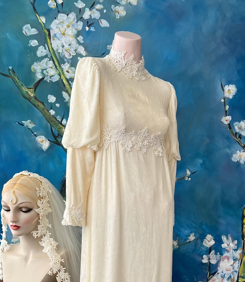 May include: Cream-colored vintage wedding dress with long sleeves and a high neckline, adorned with delicate lace detailing. The dress features a fitted bodice and a flowing skirt, displayed on a mannequin. A matching veil is also visible.