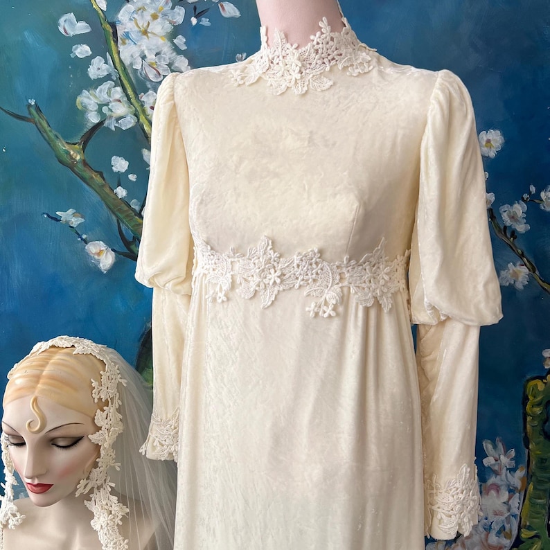 May include: Cream-colored vintage wedding dress with long sleeves and a high neckline. The dress features delicate lace detailing around the neck, waist, and cuffs. The dress has a subtle floral pattern and a flowing silhouette, evoking a sense of classic elegance.