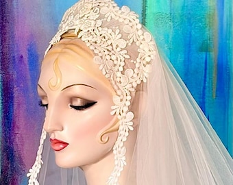 May include: An ivory lace headpiece featuring intricate floral embroidery and scalloped edges, attached to a sheer, flowing veil that drapes down the sides. The lace design covers the crown and extends along the temples in a cap-like style.