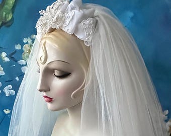 May include: A white bridal veil with a lace and fabric headband. The headband features delicate lace and fabric details. The veil is long and flowing, creating a classic bridal look. The mannequin has a fair complexion and red lipstick.