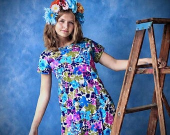 May include: A short-sleeved dress featuring a vibrant floral pattern in shades of blue, purple, green, and white. The garment is paired with a large, colorful floral crown composed of blue, orange, and white fabric flowers worn on the head.