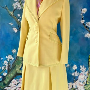 May include: A vintage yellow suit set, including a blazer and a skirt, displayed on a mannequin. The blazer features a notched collar, three buttons, and decorative stitching. The skirt is a knee-length, pleated design. A colorful scarf is visible.