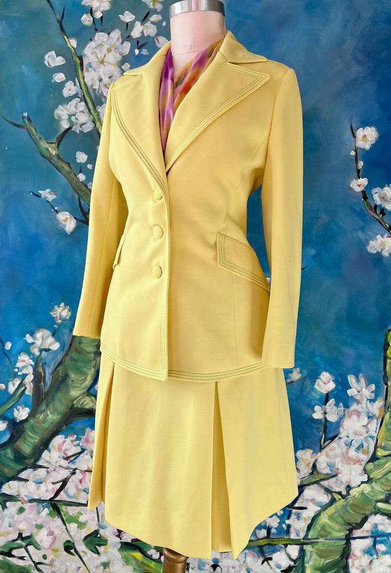 May include: A vintage yellow suit set, including a blazer and a skirt, displayed on a mannequin. The blazer features a notched collar, three buttons, and decorative stitching. The skirt is a knee-length, pleated design. A colorful scarf is visible.