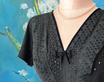 May include: A black short-sleeved dress featuring a V-neckline with a small black velvet bow at the center. The fabric is adorned with a textured, raised circular pattern and black velvet trim along the neckline. A single strand of cream-colored pearls.