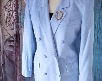 May include: A light blue double-breasted blazer featuring notched lapels, four matching buttons, and two front flap pockets. A gold-toned oval brooch with a central cameo design is pinned to the left lapel. The blazer is worn over a black top.