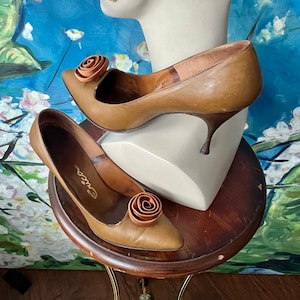 Op de afbeelding: Een paar vintage bruin lederen pumps met een rozenaccent. De schoenen hebben spitse tenen en stilettohakken. Het woord "Erica" staat op de binnenzool van één schoen. De schoenen worden op een houten standaard getoond.