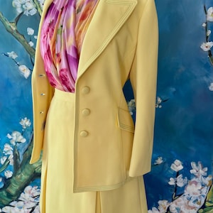 May include: A pale yellow skirt suit with a matching blazer and a pleated skirt. The blazer has three buttons and a decorative stitch. A colorful scarf with pink and purple floral patterns is draped around the mannequin's neck.